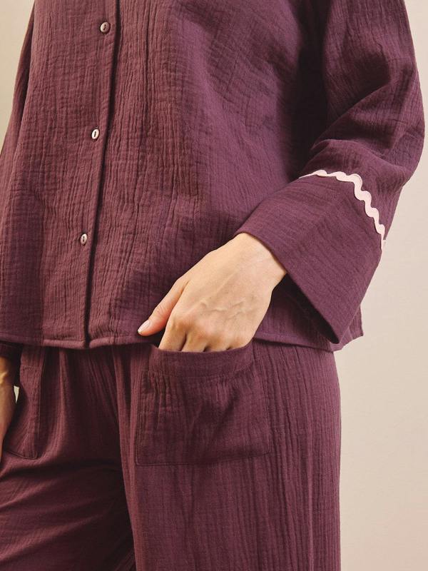 A close-up of a person wearing a matching mauve-purple crinkle gauze pyjama set with a button-front top and pants, featuring a white zigzag trim on the sleeve cuff.