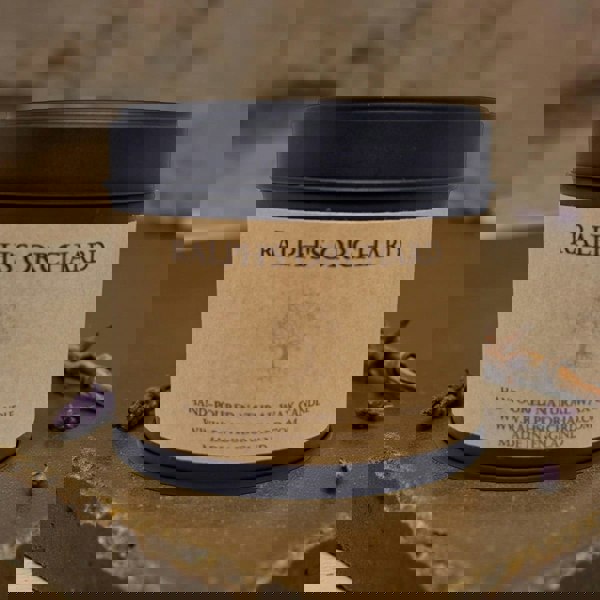 Candle in a tin labeled 'Ralph's Orchard' on a stone surface with lavender flowers.