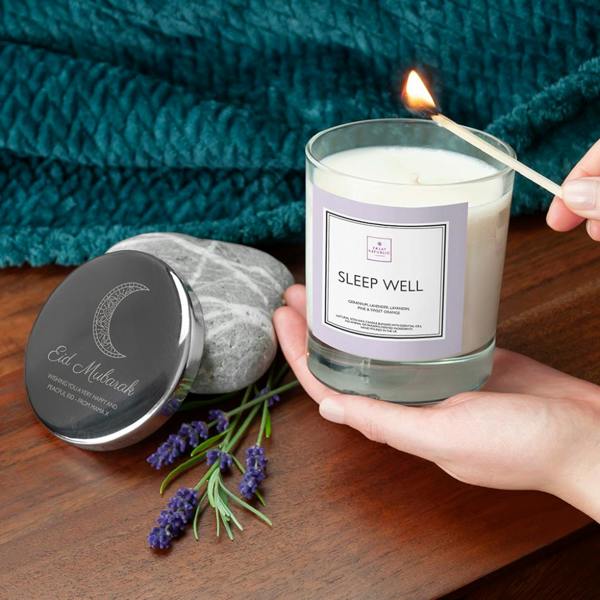 A person holds a clear glass 'Sleep Well' soy candle with a soft lilac label as it is being lit with a wooden match. Next to the candle on a wooden table sits a polished silver lid engraved with an intricate crescent moon pattern and the text 'EID MUBARAK, WISHING YOU A VERY HAPPY AND PEACEFUL EID - FROM MAMA X'. Fresh lavender sprigs, a smooth grey river stone, and a textured teal blanket complete the cozy lifestyle setting.