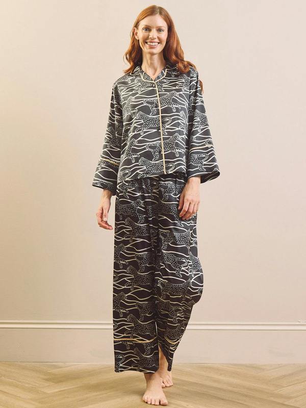 A woman stands barefoot in a studio setting wearing a matching two-piece pyjama set with a black base and an abstract wavy line pattern in white and gold including a Cheetah. The set includes a long-sleeve button-front top and matching loose pants.