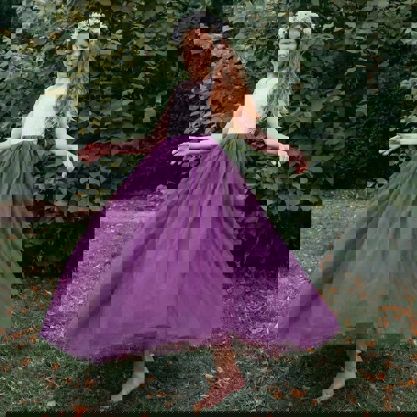 Young girl in a white top and purple skirt standing outdoors with greenery in the background