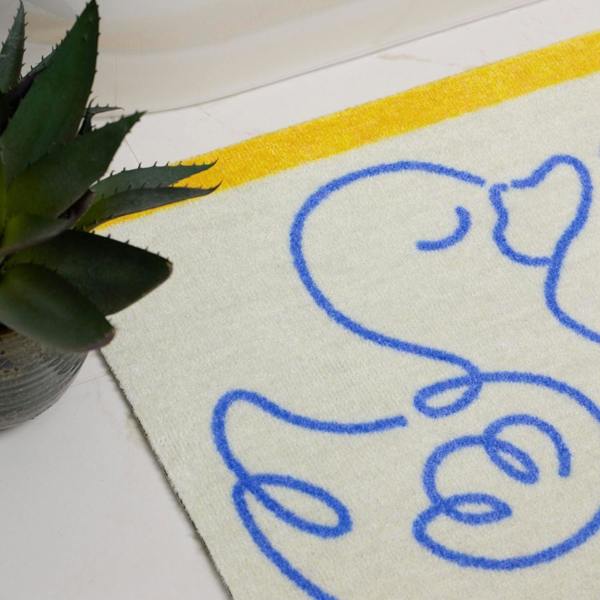 Doormat with blue duck design on a white surface next to a plant