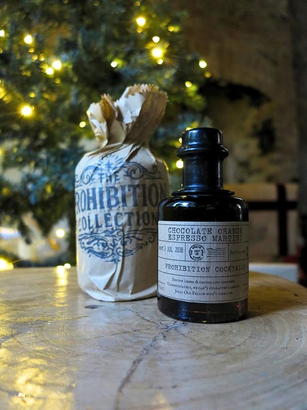 A dark glass bottle labeled 'Chocolate Orange Espresso Martini' stands on a stone surface next to a paper bag printed with 'The Prohibition Collection' and tied with twine, with a blurred festive tree of lights in the background.