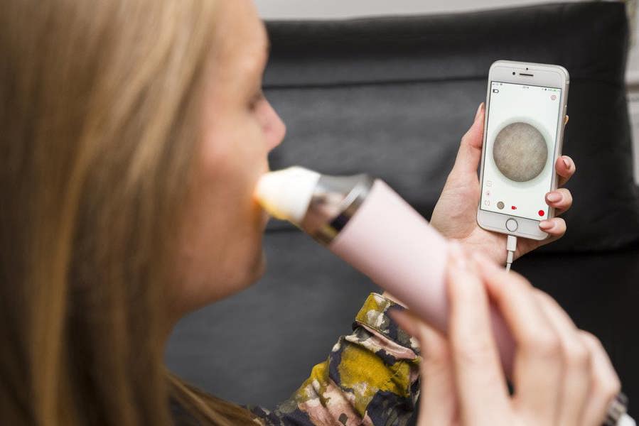 A woman uses the Rio Pore Perfection device for personalized facial skincare while viewing a magnified image of her skin on her smartphone.