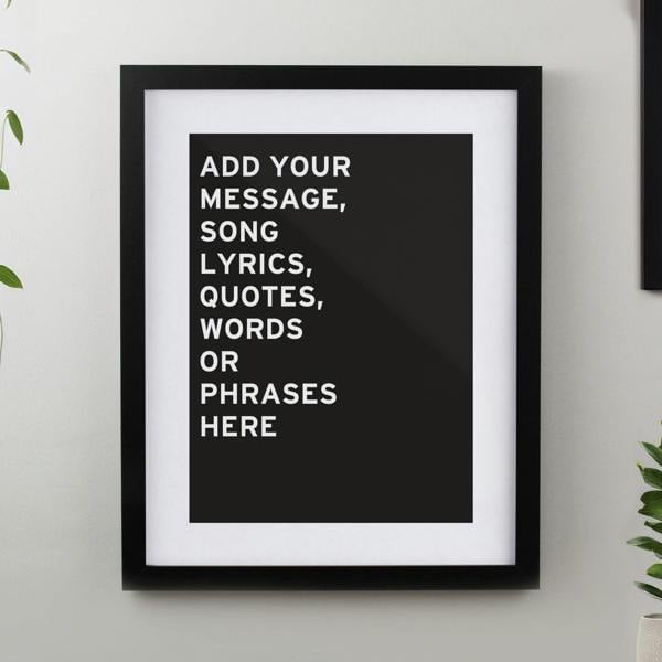 A personalised typography black framed poster print hanging on a light grey wall displaying custom white uppercase text with house plants on either side.