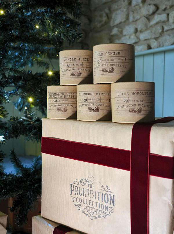 A gift set from The Prohibition Collection featuring five vintage-style paper labels for ginger and espresso martini cocktails arranged on top of a large cream-colored gift box with a dark red ribbon, set next to a decorated Christmas tree.