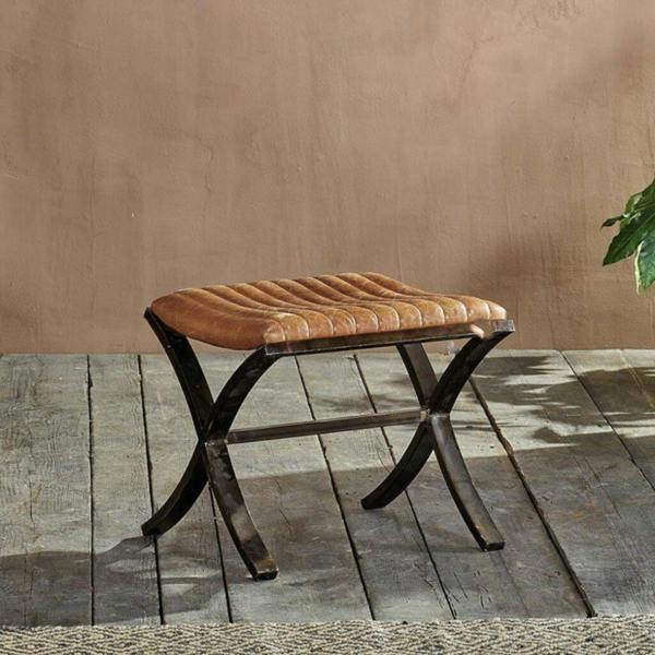 A vintage ribbed tan leather footstool with a dark metal cross-frame legs on a rustic wooden floor.