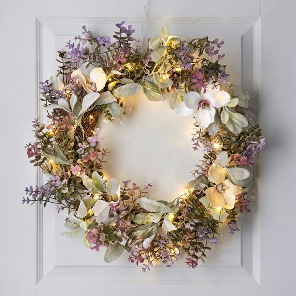 60cm oversized pre-lit spring flower wreath with warm LED lights, featuring purple eucalyptus, white magnolia blooms, and frosted green foliage hanging on a white door.