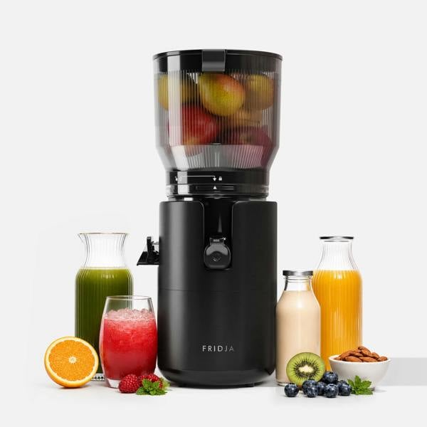 A black Fridja f2800 Open Whole Fruit Juicer filled with apples and pears is surrounded by red, green, and orange juices, a smoothie bottle, orange half, kiwi slice, raspberries, blueberries, and a bowl of almonds.