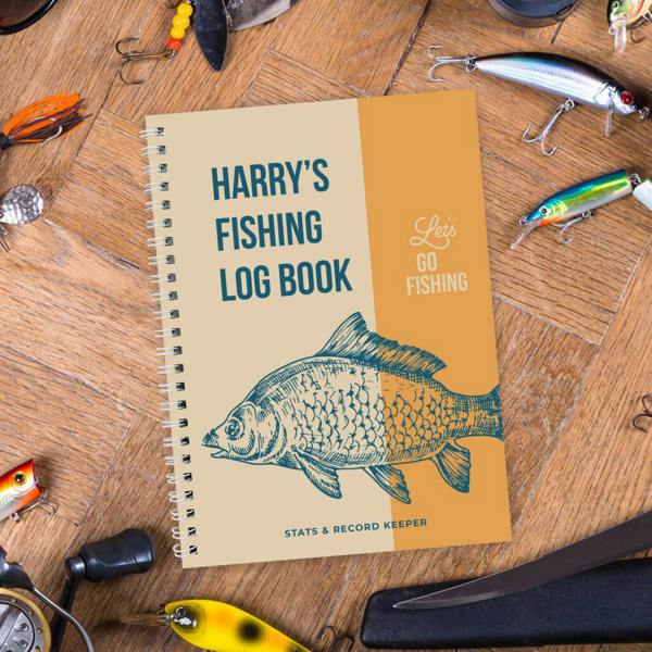 A spiral-bound Personalised Fishing Log Book featuring Harry's Fishing Log Book on a wooden table surrounded by fishing lures.