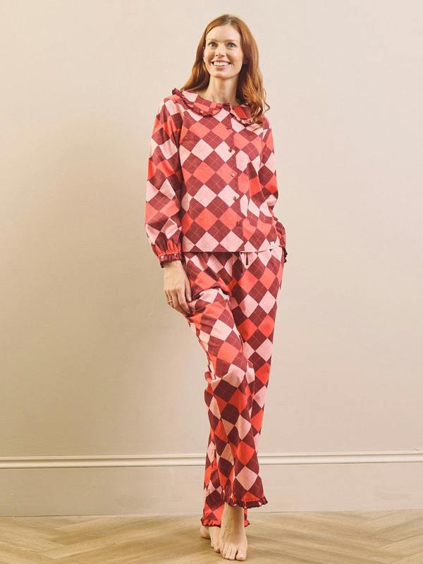 A woman modelling a matching two-piece pyjama set with a red, pink, and white harlequin diamond pattern, standing against a beige wall and light wood floor.