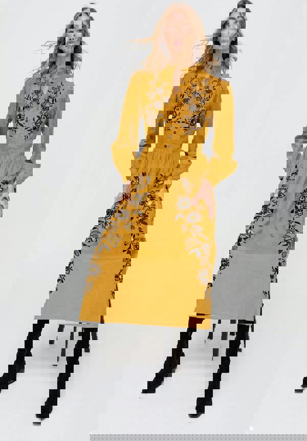 Woman wearing a mustard yellow dress with black floral embroidery on a white background