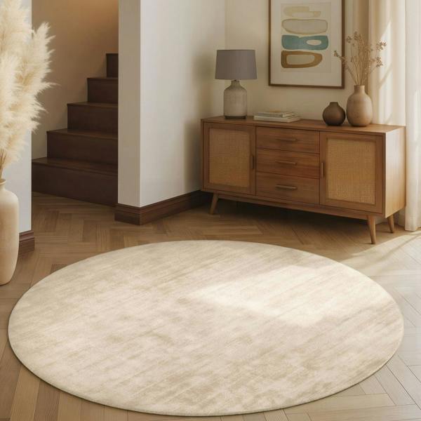 Round beige rug on a wooden floor with a wooden sideboard and decorative items in a room.
