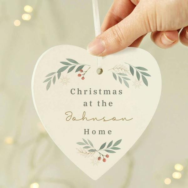 A hand holding a heart-shaped wooden Christmas tree ornament featuring a white ribbon, printed winter foliage berries, and custom family text on a cream background.