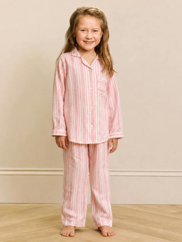 A young girl stands barefoot in a bedroom setting wearing a pink and white vertically striped pajama set consisting of a long-sleeve button-up top and matching pants.