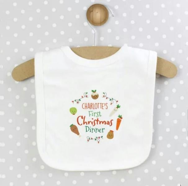 A white cotton baby bib custom printed with 'CHARLOTTE'S First Christmas Dinner' surrounded by illustrated festive food characters, hanging on a wooden hanger against a grey polka dot background.