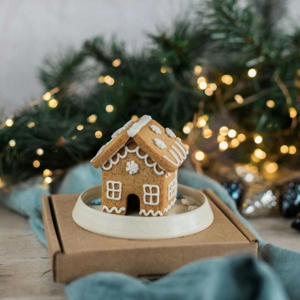 Assembled mini gingerbread house on a small ceramic dish sitting atop a kraft cardboard gift box, with fairy lights in the background.