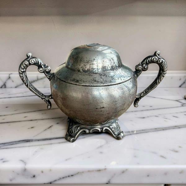 An antique silver-toned metal lidded container or caddy with ornate handles and a decorative pedestal base, placed on a white marble surface against a neutral background.
