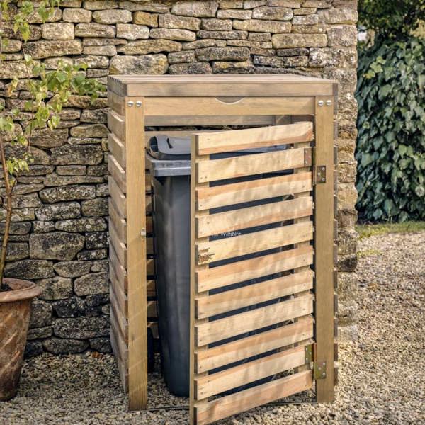 A single natural spruce timber wheelie bin store with its front slatted door slightly open, housing a grey wheelie bin on a gravel driveway by Abode Home & Gifts.