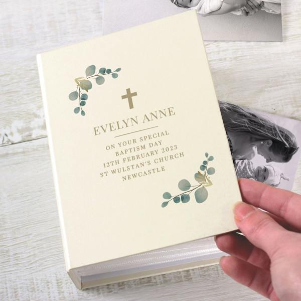 A hand holding an ivory hardback 6x4 photo album by Abode Home & Gifts next to black and white baby photos. The cover features a small cross, eucalyptus leaves, and text reading "EVELYN ANNE ON YOUR SPECIAL BAPTISM DAY 12TH FEBRUARY 2023 ST WULSTAN'S CHURCH NEWCASTLE".