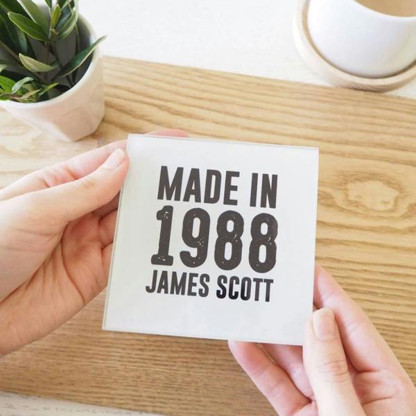 A pair of hands holding a square glass coaster that reads 'MADE IN 1988 JAMES SCOTT' over a wooden tabletop background.
