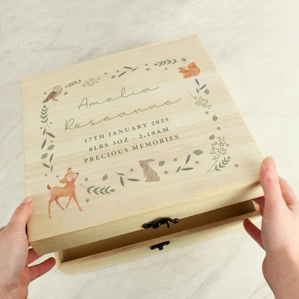 A pair of hands lifting the lid of a light wood keepsake box printed with a woodland deer, rabbit, owl, squirrel, and baby birth details.