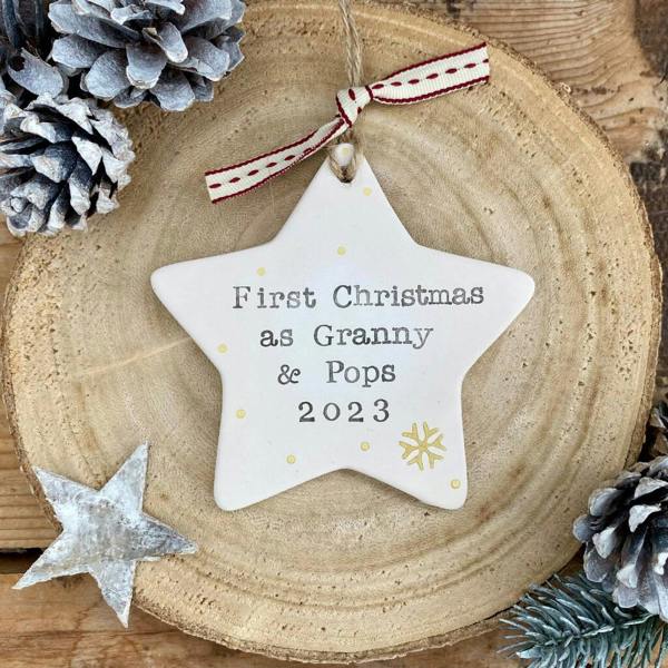 A hand-stamped white ceramic star ornament reading First Christmas as Granny and Pops 2023 with a gold snowflake on a wooden log slice.