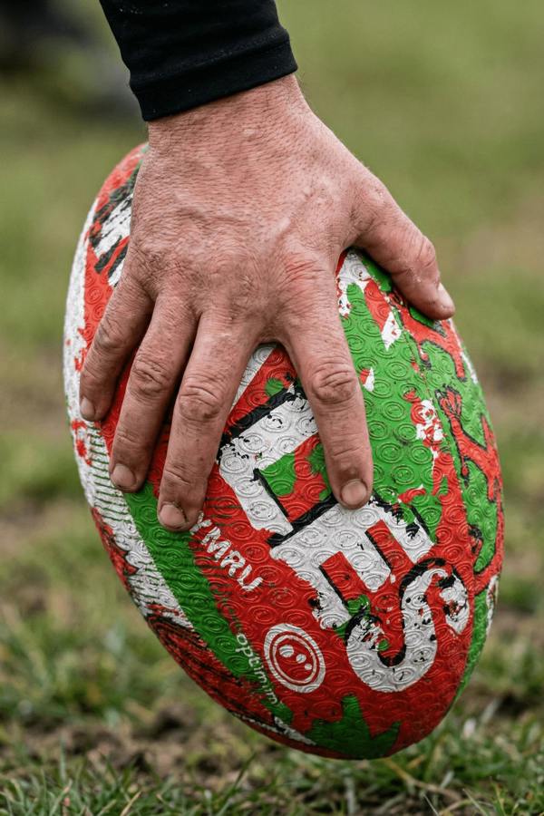 Hand holding a red, green and white Optimum Wales rugby ball with CYMRU print on muddy grass - Optimum
