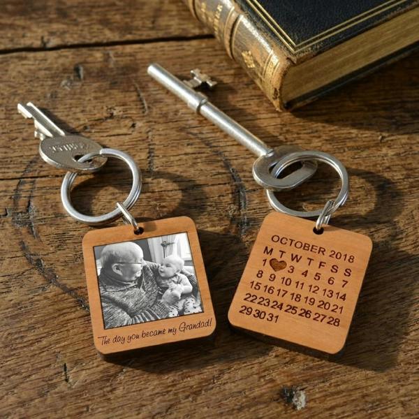 Two square cherry wood keyrings resting on a rustic old wooden table next to a vintage book and a metal key. One keyring shows a photo of a grandad and baby, the other shows an engraved October 2018 calendar with a heart marking the date.