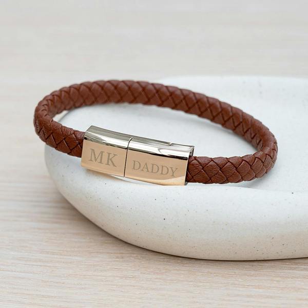 A braided tan leather men's bracelet with a polished silver magnetic clasp engraved with 'MK' and 'DADDY' resting on a small white ceramic dish.