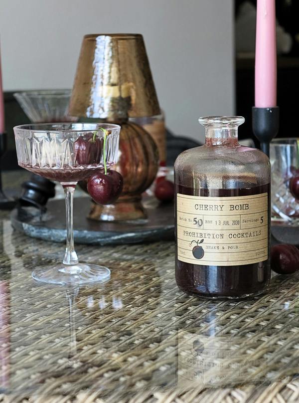 A vintage-style glass bottle labeled 'Cherry Bomb Prohibition Cocktails' filled with dark red liquid, styled on a woven table with a coupe glass, cherries, and a metallic container in the background.