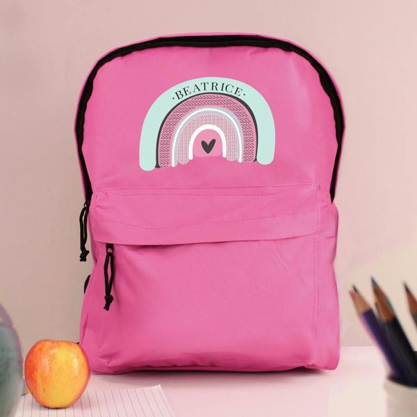 A personalised pink backpack featuring a boho pastel rainbow design and printed with the name Beatrice, styled on a desk alongside an apple and coloured pencils.
