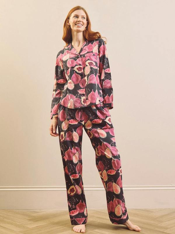 A model wearing a matching pyjama set with a pink and red fig pattern on a black background, set against a neutral beige wall and light wood floor.