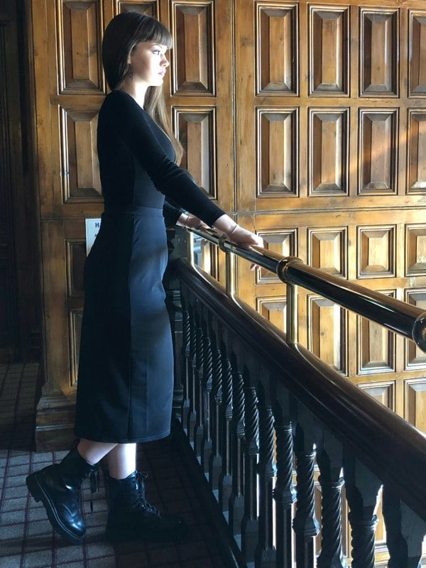 This is a young woman wearing a black long sleeve t-shirt with a below the knee, pencil style skirt, with a pleated waist. She has long brown hair and is wearing shiny black boots. She is silhouetted against a dark wood panelled room, leant against a banister, overlooking the room below.