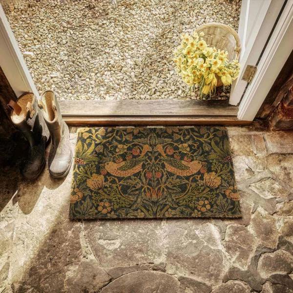 Decorative doormat with floral pattern on stone floor, shoes to the left, basket with flowers above.