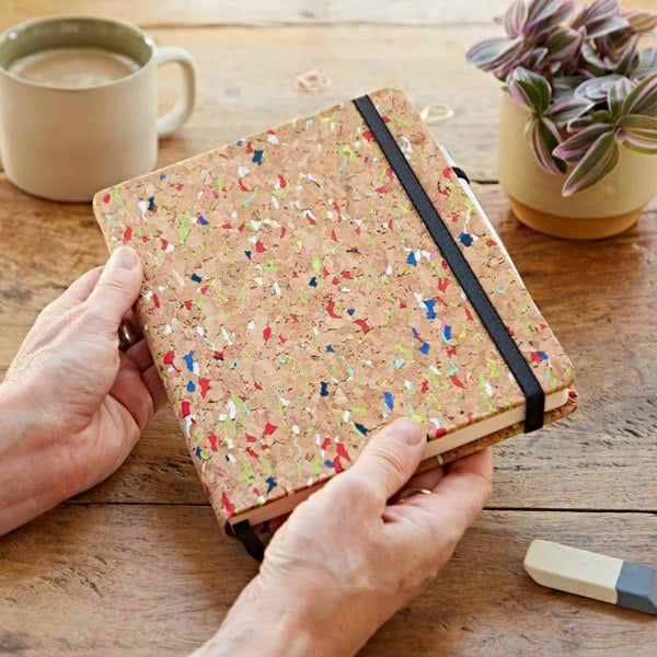 A person holding a Natural Cork A5 Notebook on a wooden table.