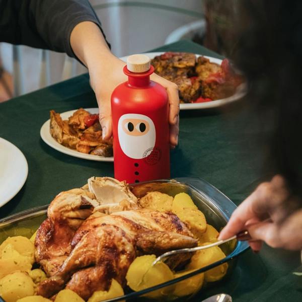 Person reaching for the 500ml Santa edition extra virgin olive oil bottle at a Christmas dinner table surrounded by roast chicken and potatoes.