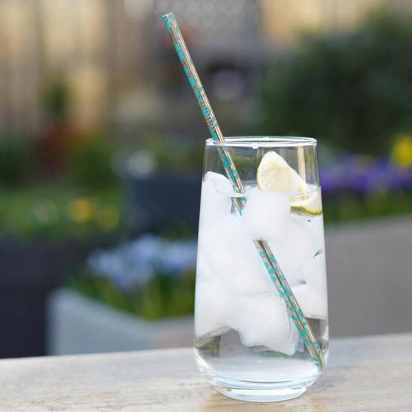 A stainless steel straw with a turquoise floral pattern inside a clear glass filled with water, ice cubes, and a fresh lime wedge.