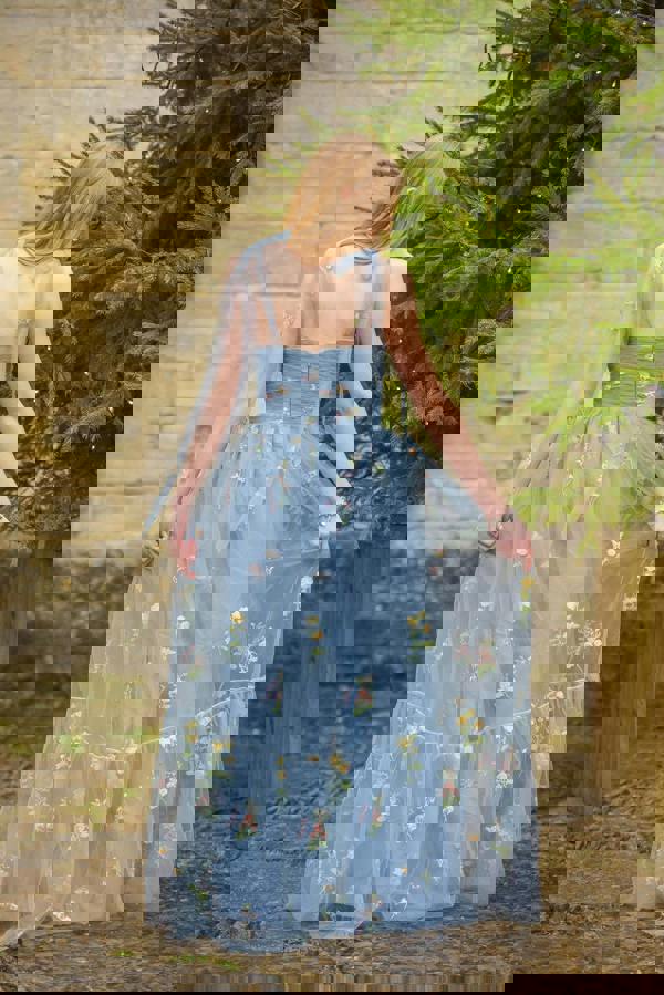 Woman in a light blue floral dress standing outdoors against a stone wall and greenery.
