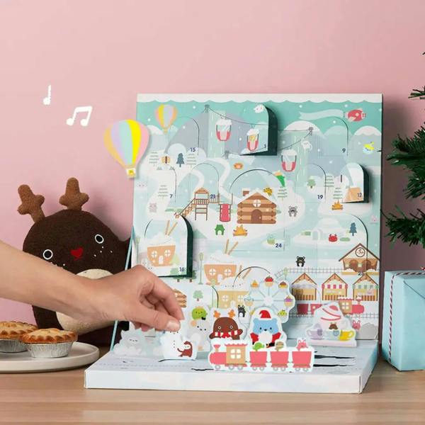 A hand opening a window on a pastel-colored musical Christmas advent calendar featuring cartoon characters on a ski slope and magnetic characters on a skating rink.