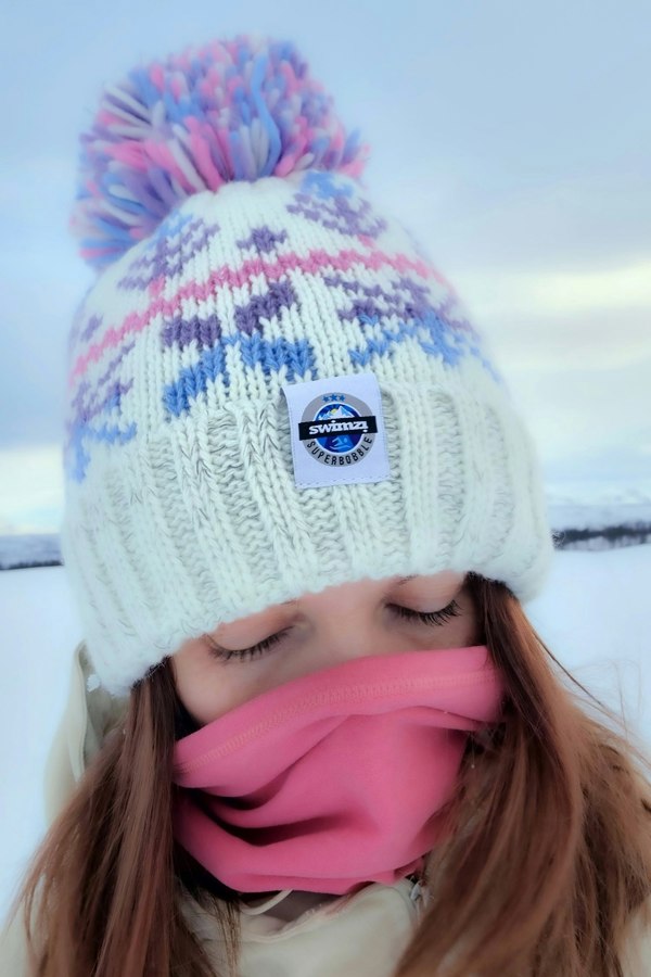  Woman in the snowy foothills of Northern Norway is wearing a chunky Nordic knit bobble hat. The white base and cool pastel toned Nordic design hat also has a deep ribbed headband, contains reflective yarns and has a luxury sherpa fleece lining.