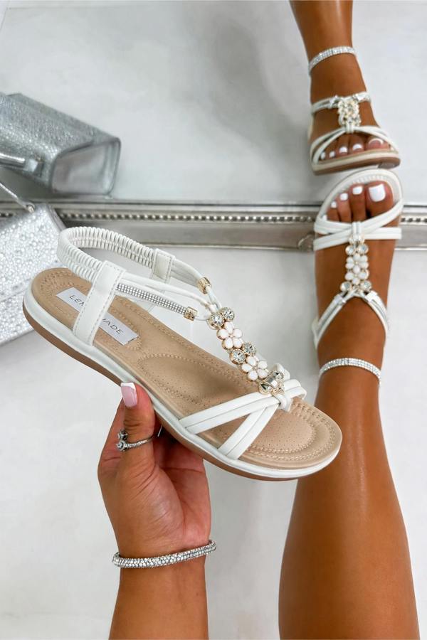 White strappy flat sandals with pearl and crystal embellishments, shown being worn on feet and held in a hand against a light gray background.