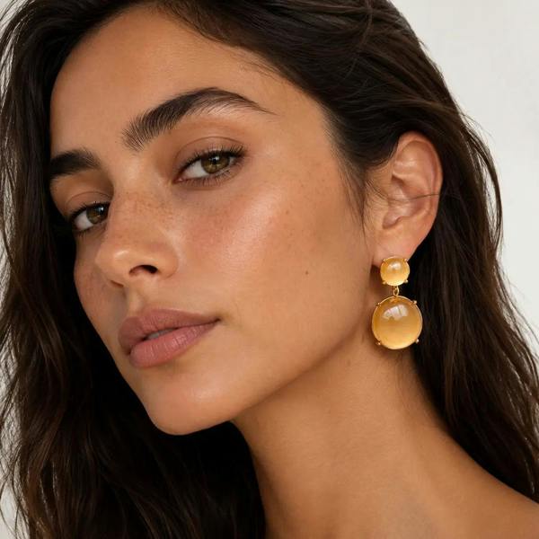 A close-up of a woman wearing a pair of chunky, double-drop dangle earrings made of warm yellow-gold-toned stone and metal, set against a clean, light off-white background.