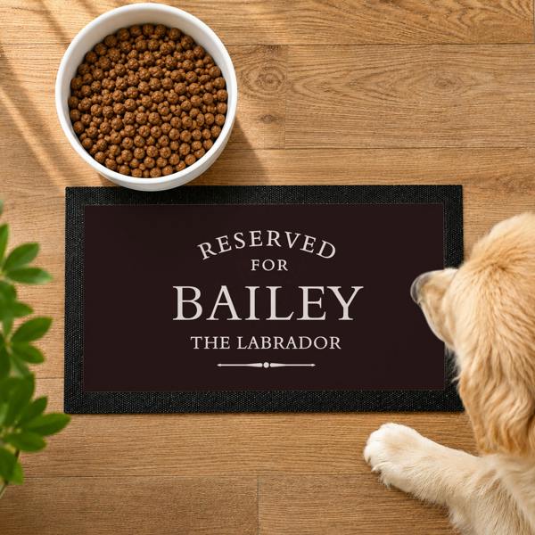 Top-down view of a dark personalised pet bowl mat reading RESERVED FOR BAILEY THE LABRADOR next to a dog and a food bowl.