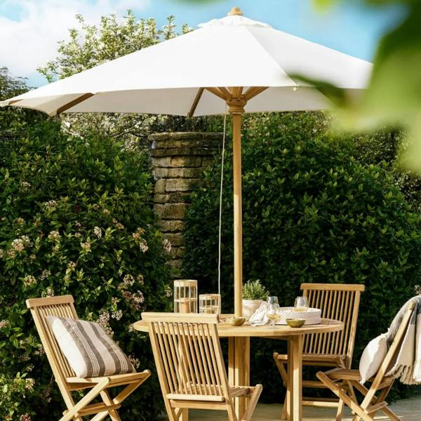 A 3m natural cream canopy teak wooden parasol shading a round wooden dining table and four matching chairs on a paved garden terrace.