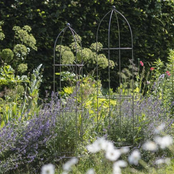 A pair of raw metal garden obelisks of different heights positioned among purple flowers and green shrubs.