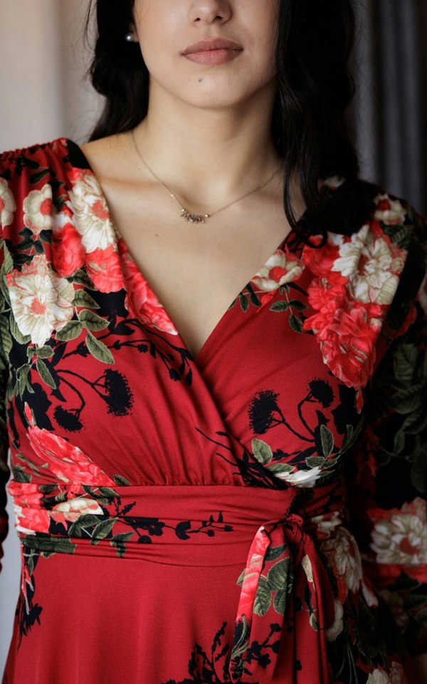 This is a close up of a young lady wearing a V neck style mock wrap dress, with a gathered bust and shoulders and a tie at the waist. The dress is a red floral print. The model has dark hair, worn behind her shoulders