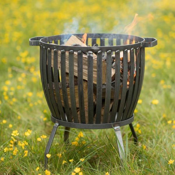 The small Barrington Fire Pit crafted from raw metal strapping, shown lit with burning logs in a wildflower meadow.