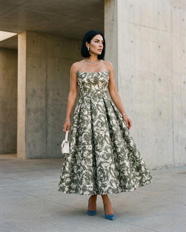 A woman stands outdoors against a concrete wall, wearing the Sarvin Tala Corset Midi Dress, styled with straight black hair, jewelry, blue high heels, and holding a small white purse.