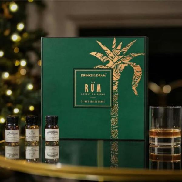 A luxury rum advent calendar in a green and gold box on a glass table next to wax-sealed miniatures and a glass of rum.